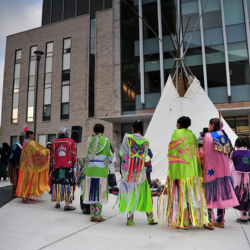 Indigenous community members in regalia with a teepee on front of a campus building.
