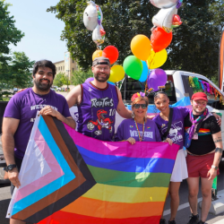 Western Pride parade with people holding pride flag. Balloons and decor in background.
